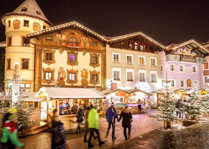 Abendlicher Blick auf den Marktplatz von Berchtesgaden. Im Vordergrund verschneite Bäume und die weihnachtlichen Stände vom Weihnachtsmarkt