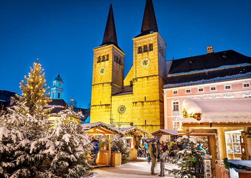 Abendlicher Blick auf das beleuchtete Schloss von Berchtesgaden. Im Vordergrund verschneite Bäume und die weihnachtlichen Stände vom Weihnachtsmarkt