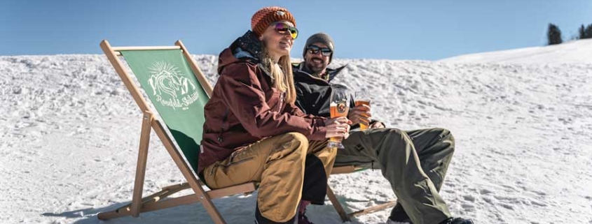 Sonniges Wetter und Schnee - Entspanntes Pärchen sitzt im Liegestuhl und trinkt ein Weissbier