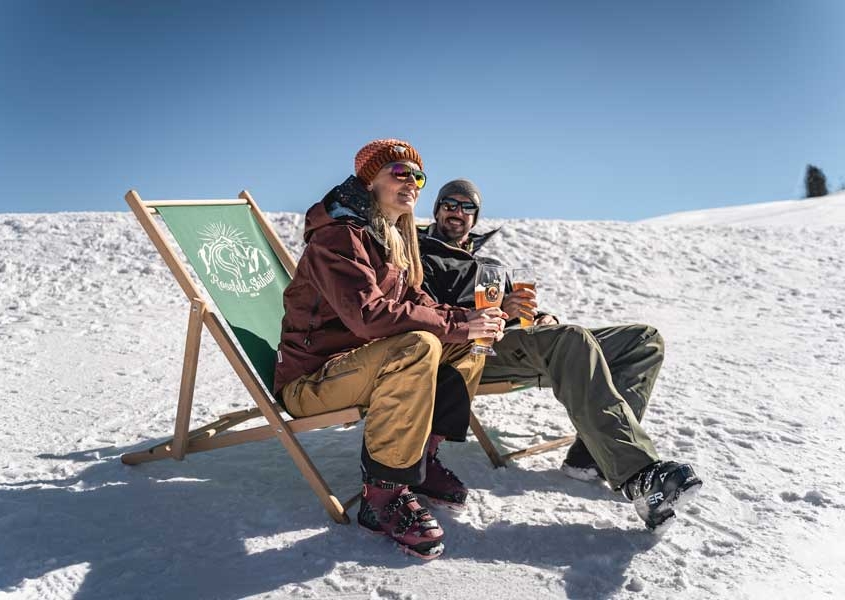 Sonniges Wetter und Schnee - Entspanntes Pärchen sitzt im Liegestuhl und trinkt ein Weissbier
